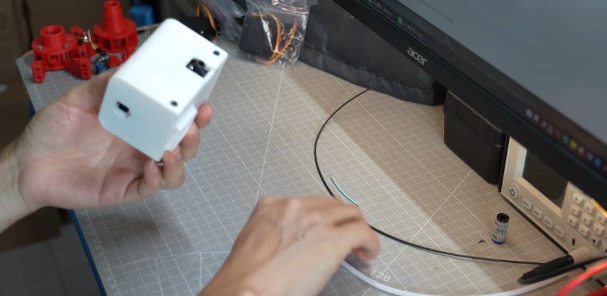 Hand holding a Bambu Lab TPU Feed Assist Module on a maker’s desk workbench background